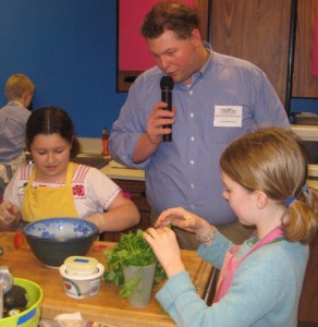 Zora and Arla prepare their winning A to Z Carnitas while event MC, Kyle Freeman, shares what they are doing with the audience.