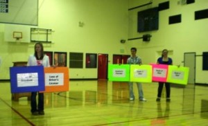 Point Blank students holding cards showing the consequences of "a little party with some friends." Note the "parents" on the left, holding three of the four cards indicating the number of times they were sued.