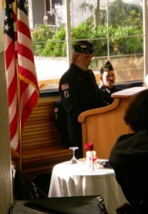 Larry Everett at the podium at the Orcas High School Commons, with George Jacson seated behind him, and "The Table for One" before him.